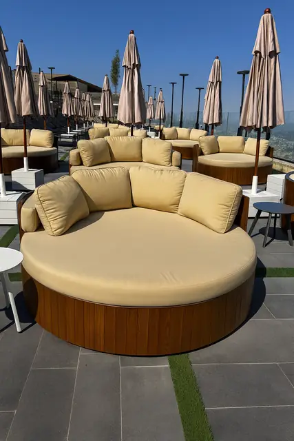 A round poolside daybed with a slatted oak base and pale cushions.